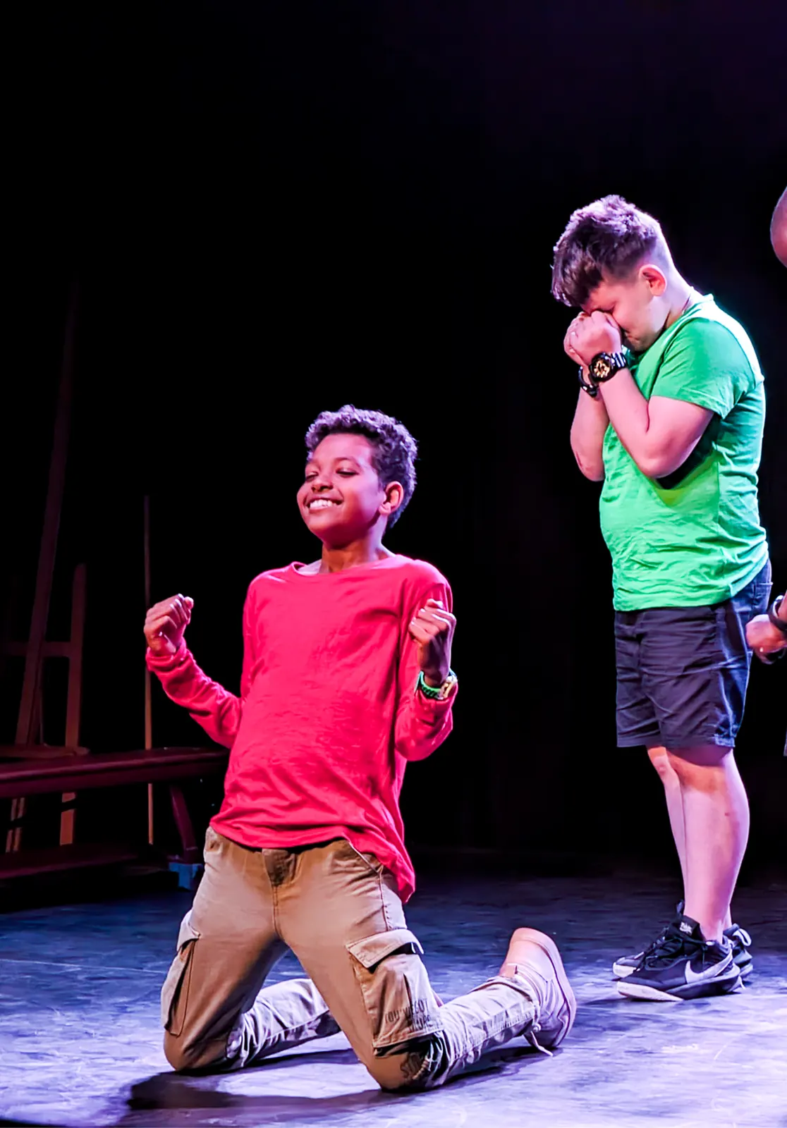 Een jongen in een rood shirt viert op zijn knieën op het podium terwijl een medespeler emotioneel toekijkt