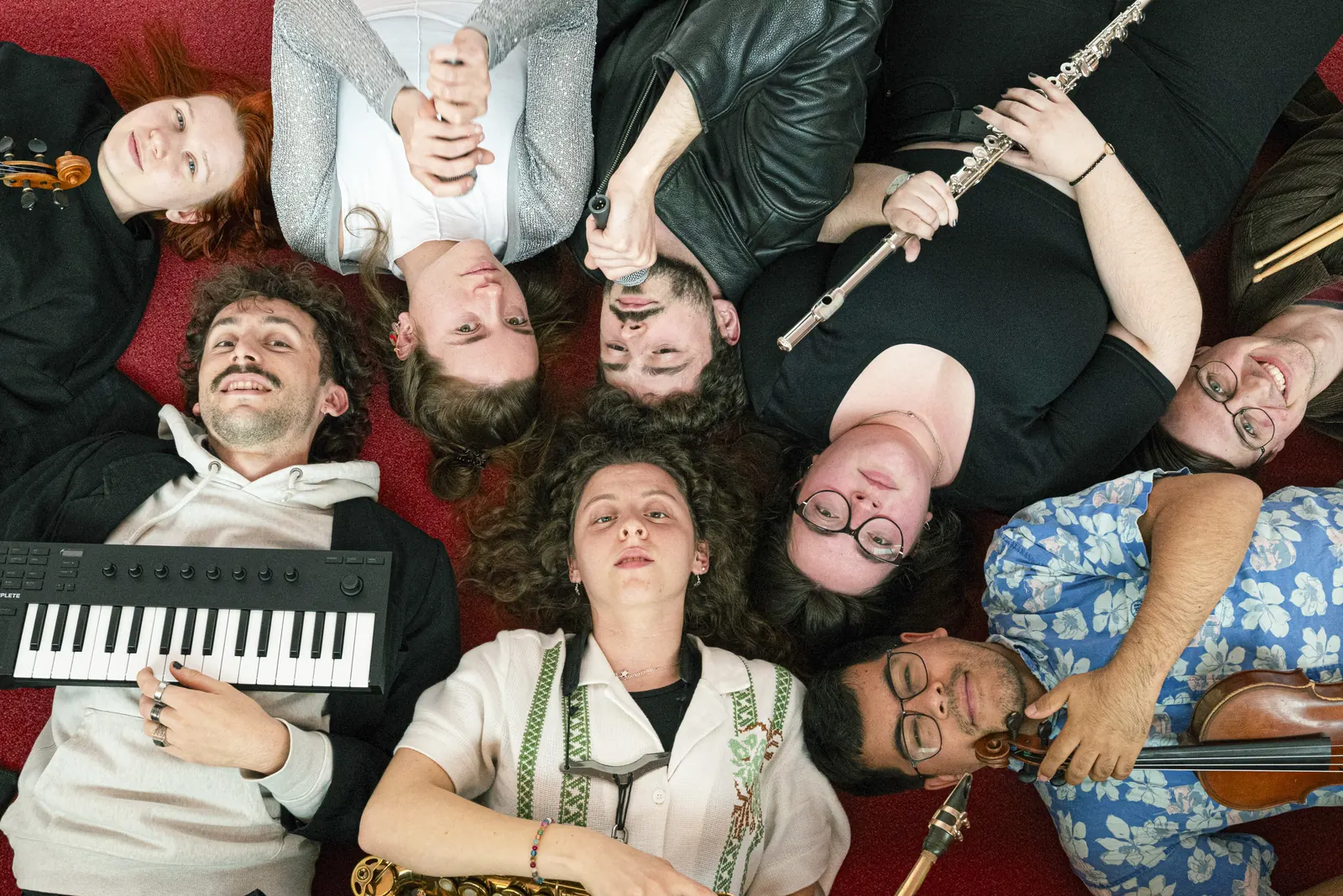 Muzikanten van Maisha in Concert liggen in een kring op een rood tapijt, met keyboard, fluit, saxofoon en viool